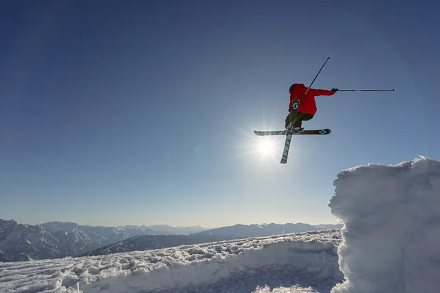 Freeskier in roter Jacke springt mit gekreuzten Skiern vor der Sonne über eine Schneewechte